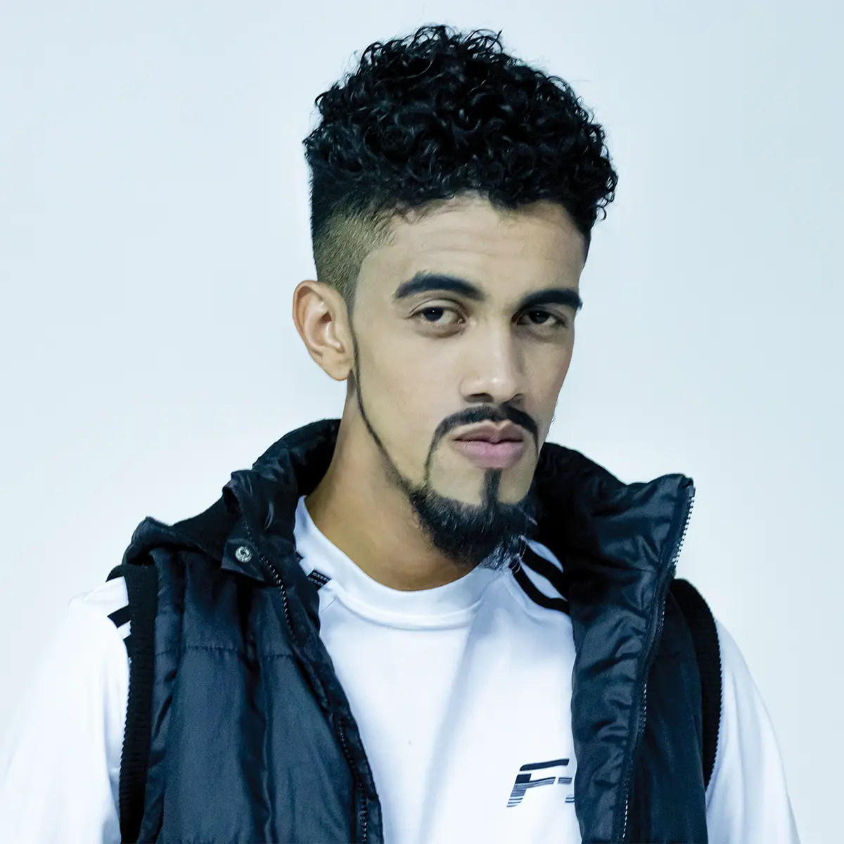 Retrato de um jovem de expressão séria, com cabelo cacheado e bem definido, barba estilizada, vestindo camiseta branca e colete preto. Fundo branco e iluminação suave.
