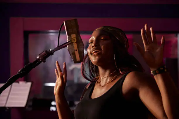African-american female singer recording r&b vocals on microphone in professional music studio recording booth