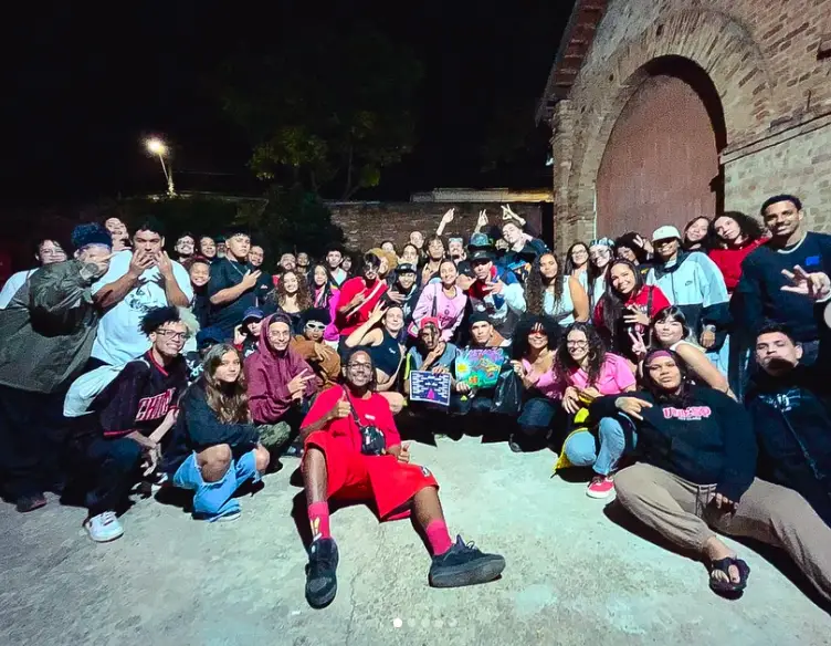 Foto noturna de um grande grupo de pessoas reunidas em frente a uma construção de tijolos à vista, sorrindo e fazendo sinais com as mãos. A maioria é jovem, com roupas urbanas e estilos diversos. No centro, pessoas seguram placas coloridas da Batalha Estação da Rima. O ambiente transmite união, celebração e energia cultural periférica.