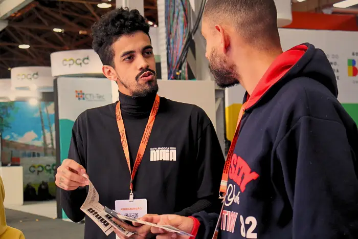 Duas pessoas conversam em pé durante um evento, um deles veste camiseta da Catalisadora Maia e segura materiais informativos enquanto explica algo ao outro participante.