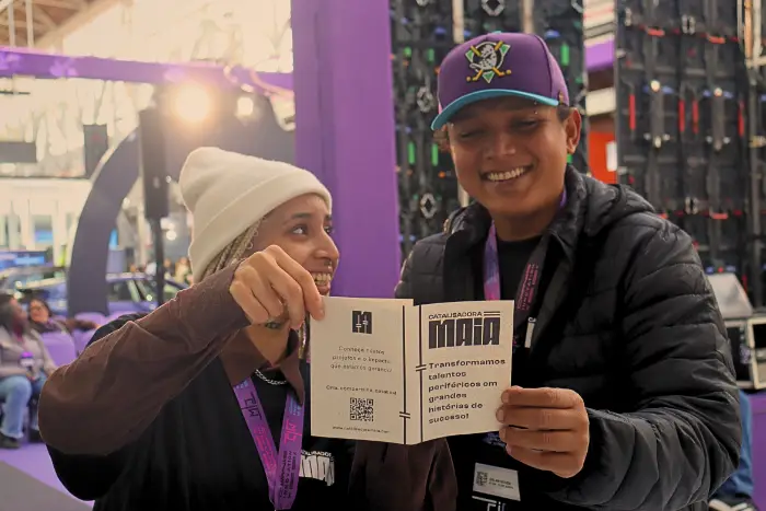 Duas pessoas em evento cultural seguram um material impresso da Catalisadora Maia e sorriem enquanto conversam, em um espaço de feira com estrutura tecnológica ao fundo.