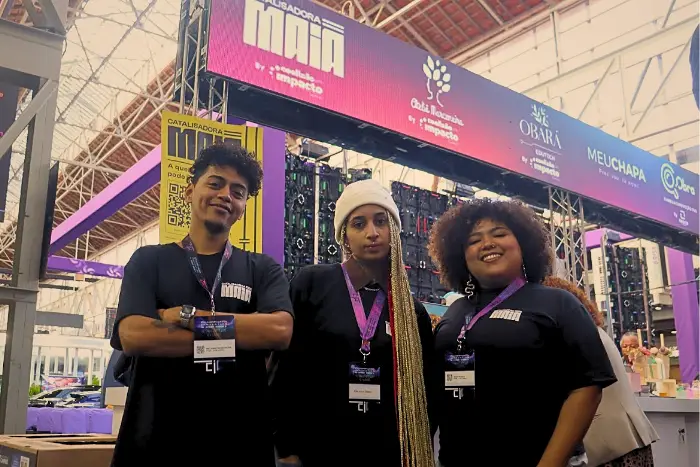 Três pessoas posam lado a lado usando camisetas da Catalisadora Maia em um estande de evento, com estrutura de palco e painéis de marcas ao fundo.