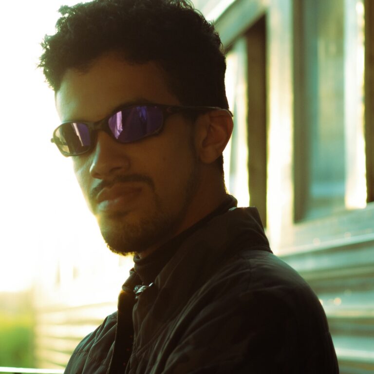 Retrato de homem usando óculos escuros e jaqueta preta, iluminado pela luz do fim de tarde em ambiente urbano.